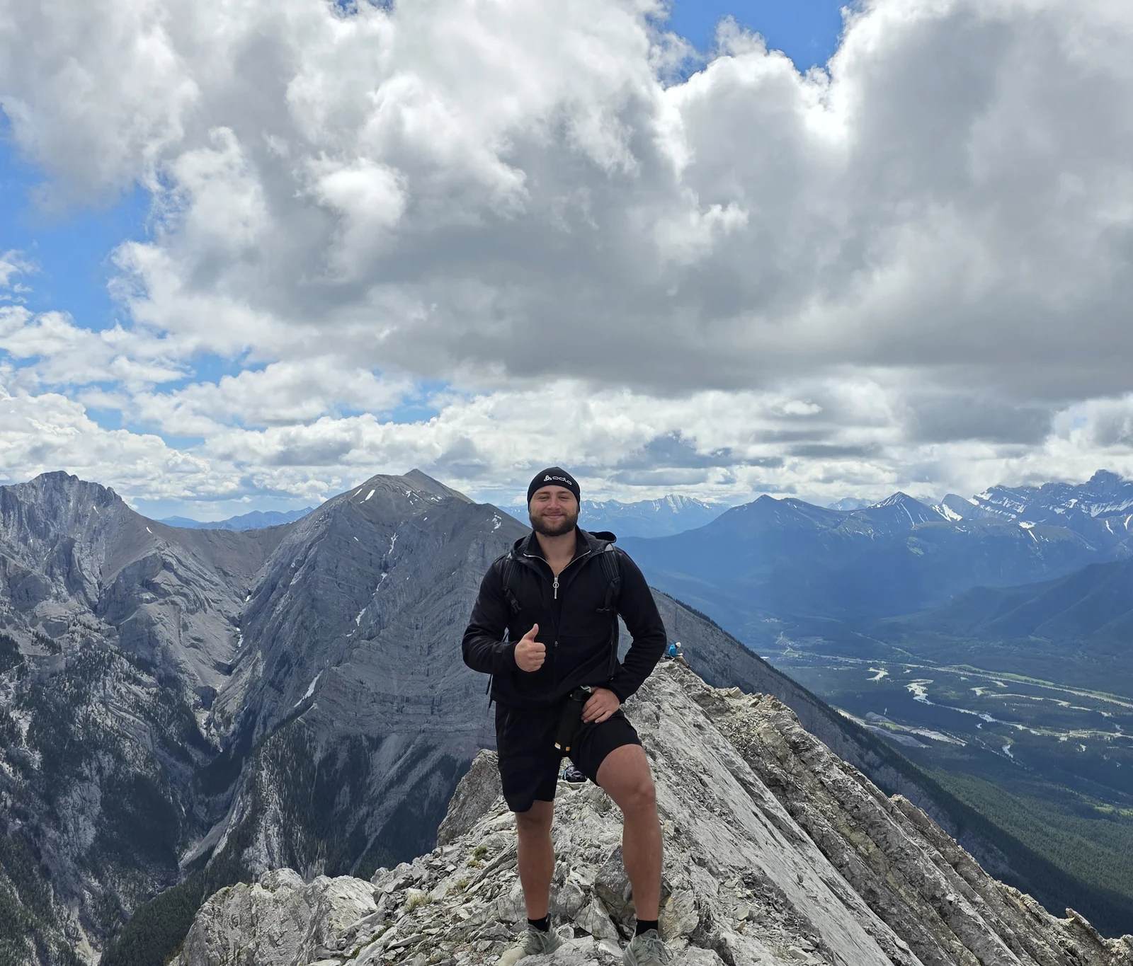 Toni auf einem felsigen Berggrat, im Hintergrund Gipfel, Wolken und ein weites Tal.
