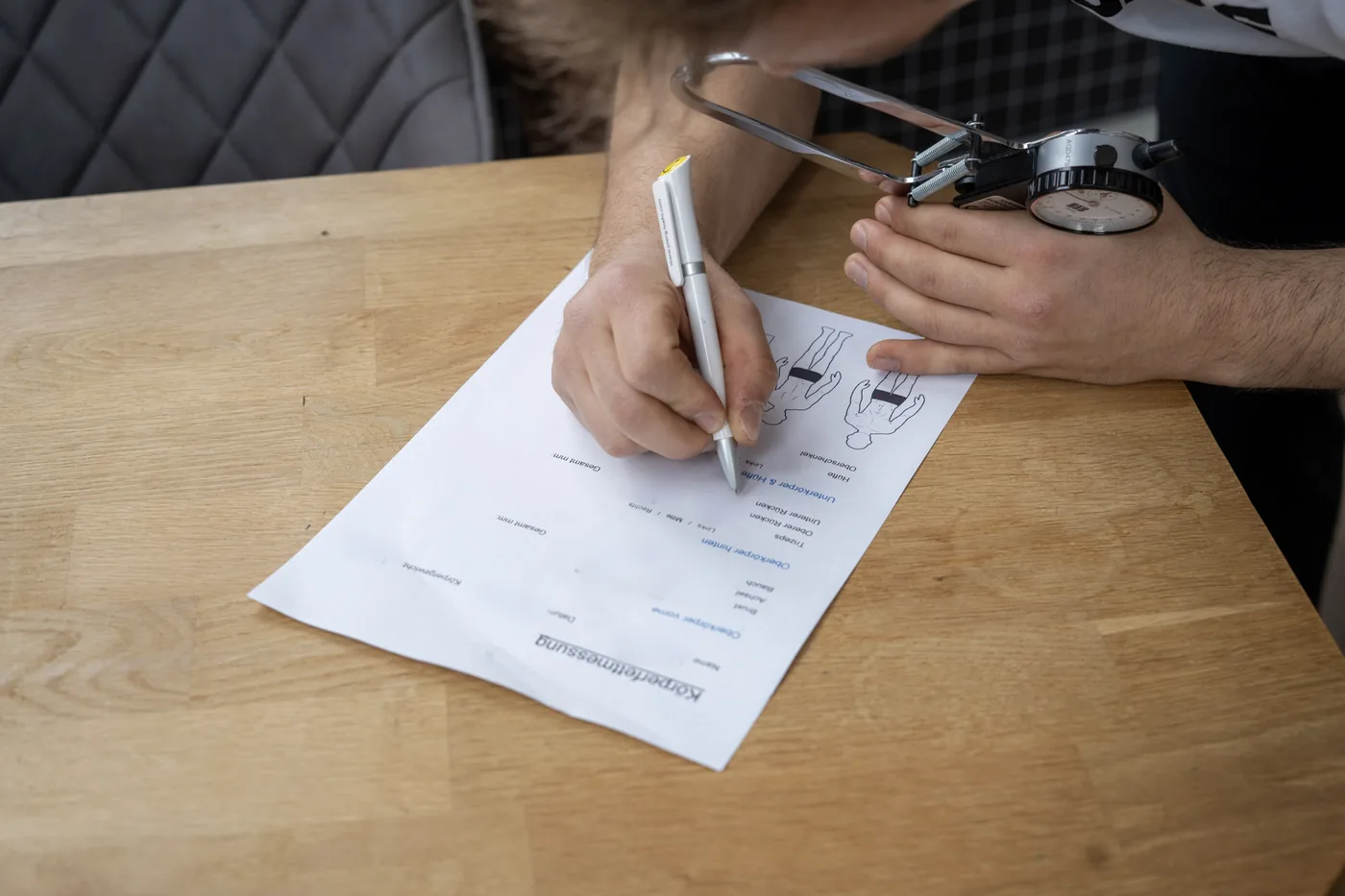 Messwerte werden mit Kugelschreiber in einen Messbogen zur Körperfettmessung eingetragen, daneben das Hautfaltenmessgerät. Name und Messwerte sind unkenntlich gemacht.