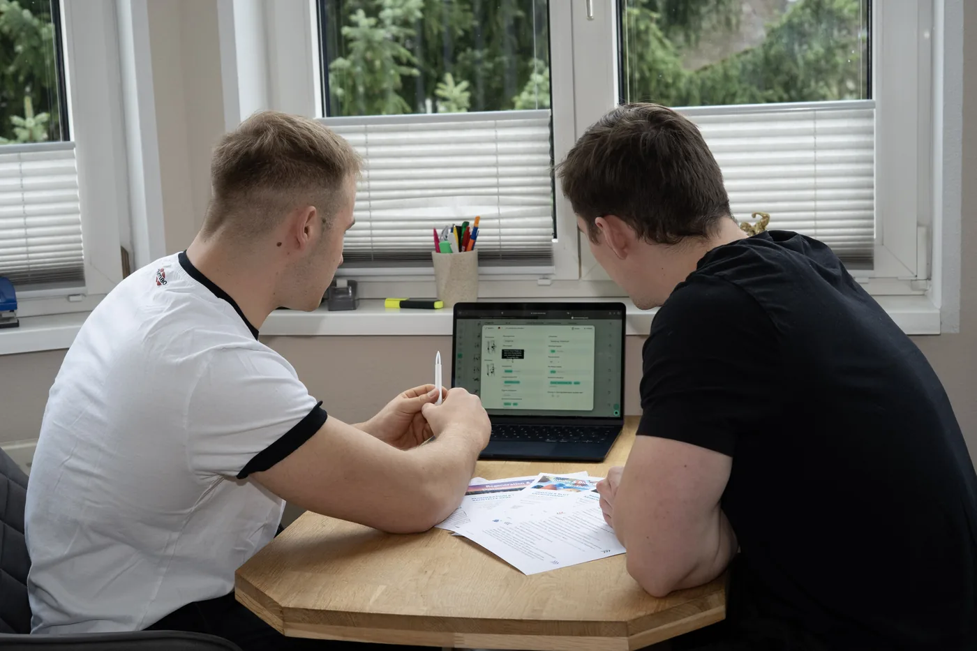 Toni und ein Kunde sitzen am Tisch und sehen sich gemeinsam eine Auswertung am Laptop an, daneben ausgedruckte Unterlagen.
